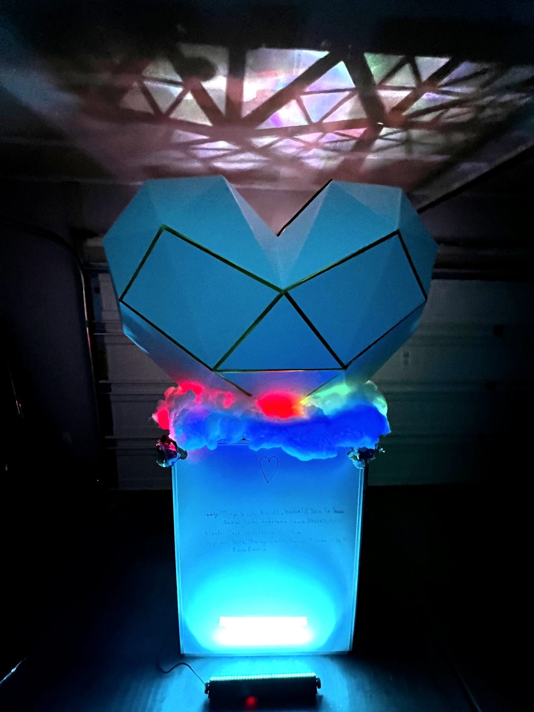 The full concept mocked up at scale at night: glowing heart, rainbow-lit cloud, handwritten names on the pedestal
